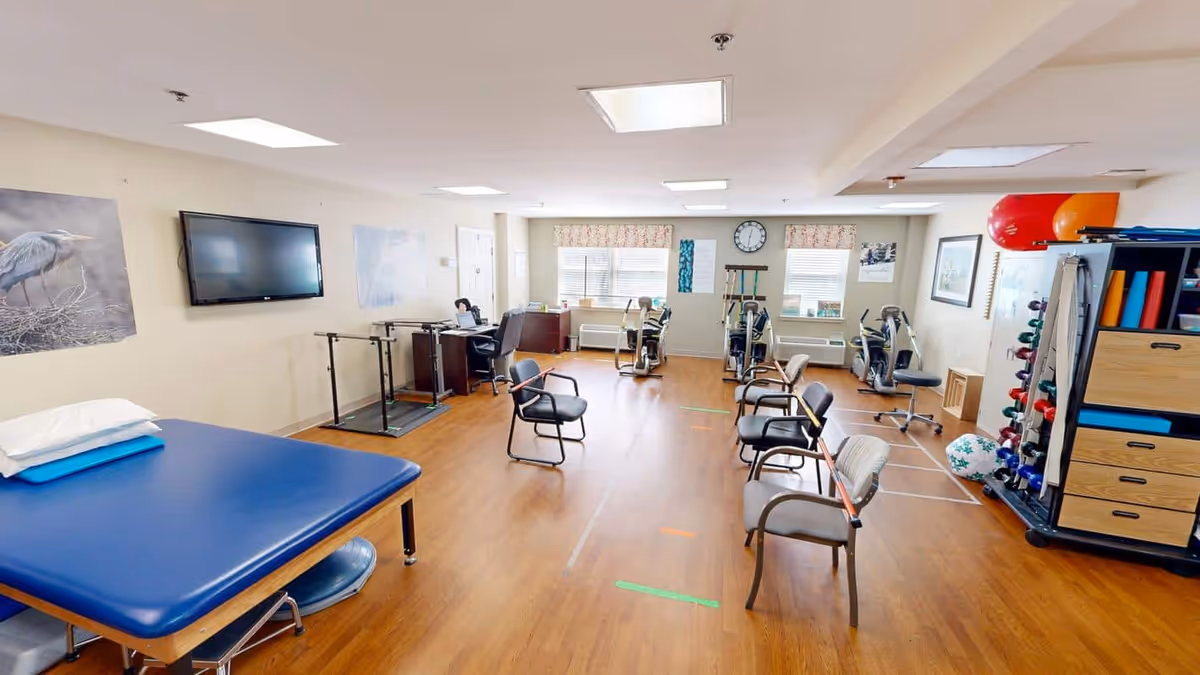 A spacious physical therapy or rehabilitation room with wooden flooring, several chairs arranged in the center, exercise equipment including stationary bikes, parallel bars, and a blue padded therapy table with pillows. The room has bright lighting from ceiling fixtures and windows with floral curtains. There are exercise balls, weights, and storage cabinets along the walls, with nature-themed artwork and a clock on the far wall.