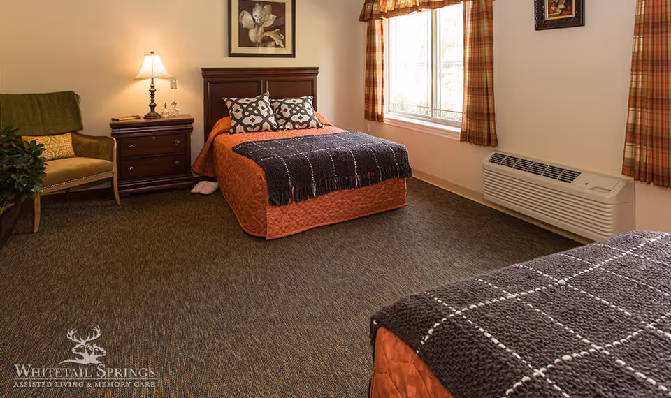 A cozy bedroom in Whitetail Springs Memory Care Community featuring two beds with orange bedspreads and dark gray knitted throws, a wooden nightstand with a lamp, a green armchair, a window with plaid curtains, and framed artwork on the walls.