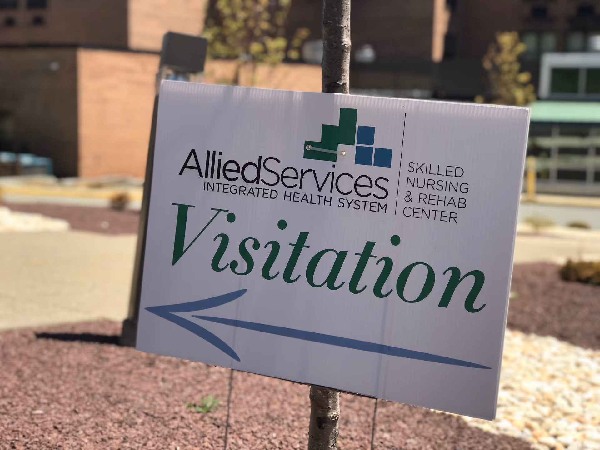 A white sign attached to a tree with the Allied Services Integrated Health System logo and text indicating 'Skilled Nursing & Rehab Center'. The sign has the word 'Visitation' in large green letters with a blue arrow pointing left. The background shows an outdoor area with mulch, rocks, and a building.