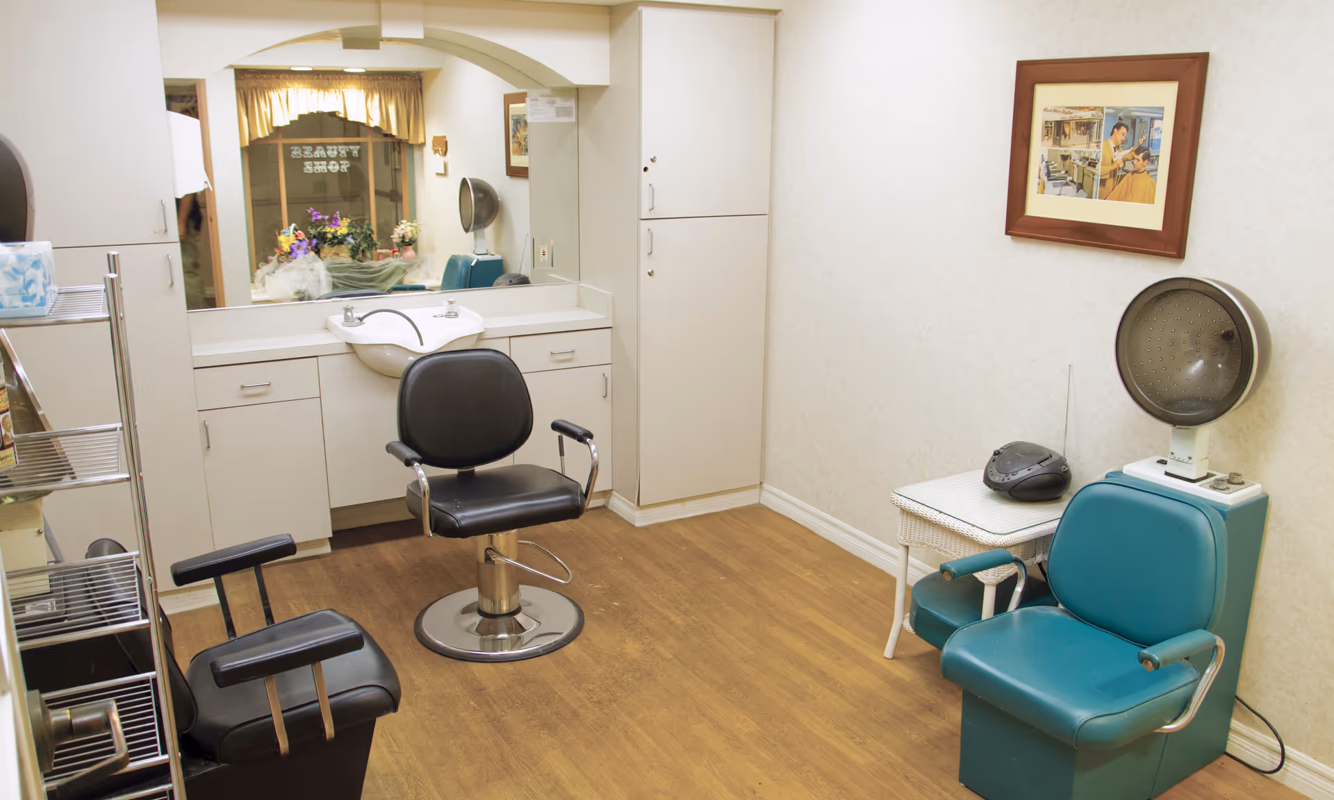 Small beauty salon room with styling chairs, a wash basin, hooded dryer, mirror, and cabinets.