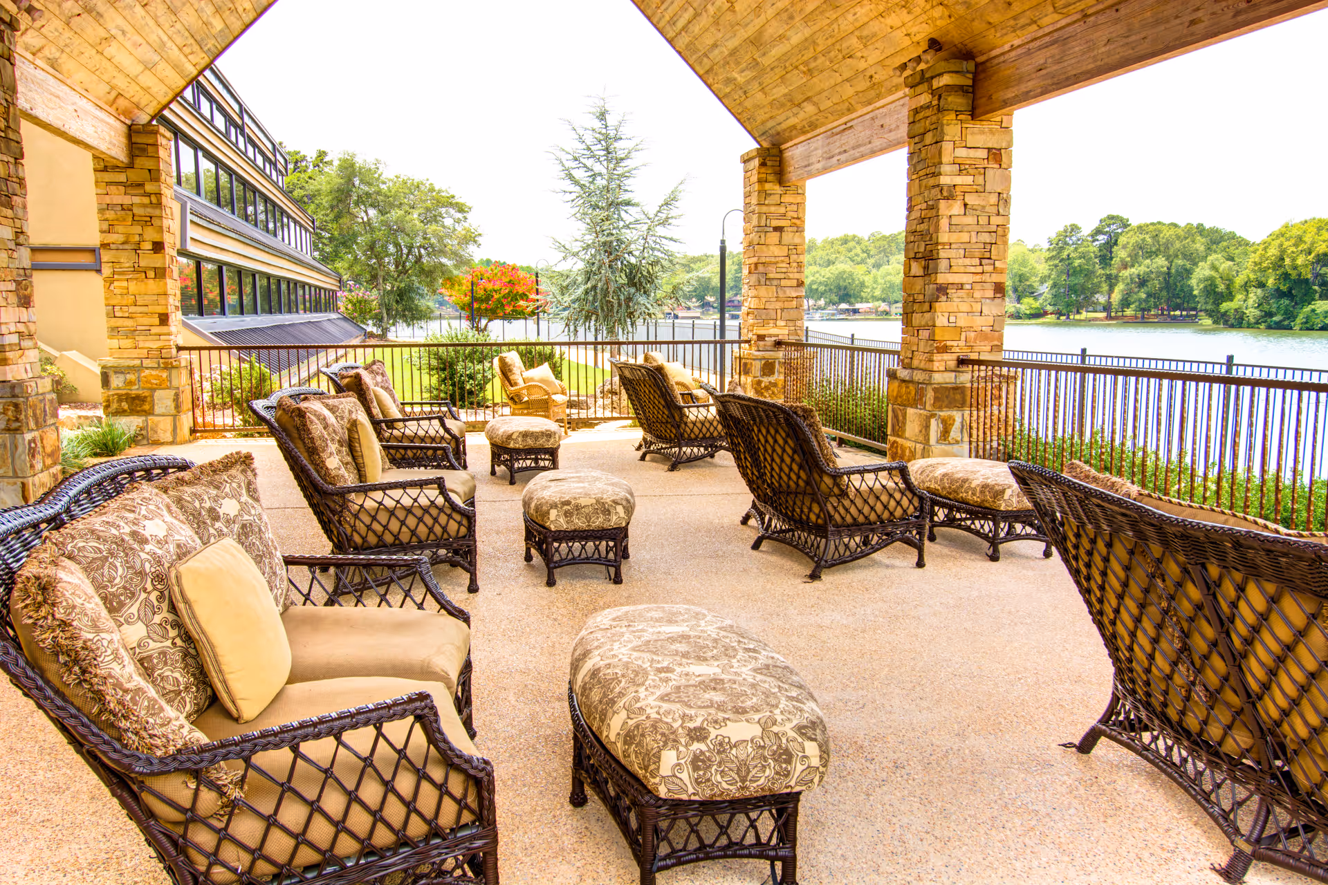 Covered outdoor patio area with multiple cushioned wicker chairs and ottomans arranged facing a scenic view of a lake and surrounding trees. The patio has stone pillars and a wooden ceiling.