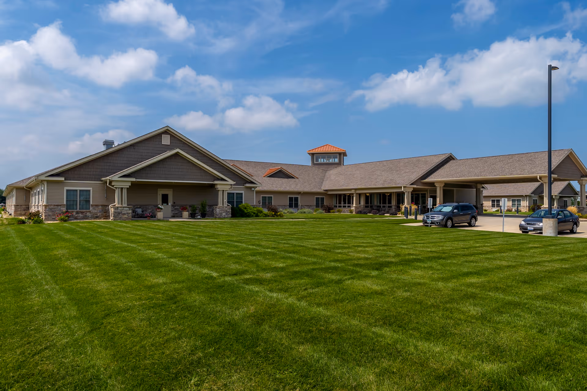Exterior view of The Carriages of Bloomington senior living facility showing a large, single-story building with a covered entrance, several windows, and a well-maintained green lawn under a partly cloudy blue sky. Two cars are parked near the entrance.