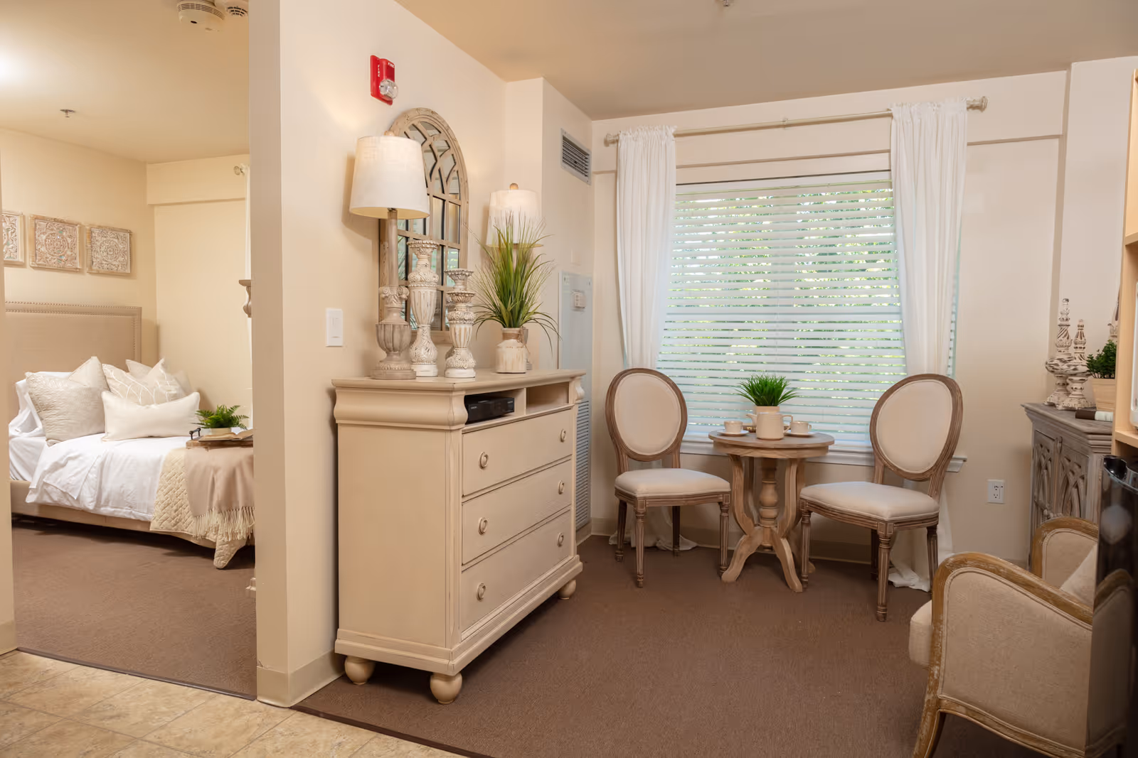 A cozy assisted living facility room with a small sitting area featuring two upholstered chairs and a round wooden table with a potted plant and cups. To the left, there is a dresser with decorative lamps, a mirror, and plants. In the background, a bedroom with a neatly made bed and pillows is visible. The room has beige walls, carpeted flooring, and a window with white curtains and blinds.