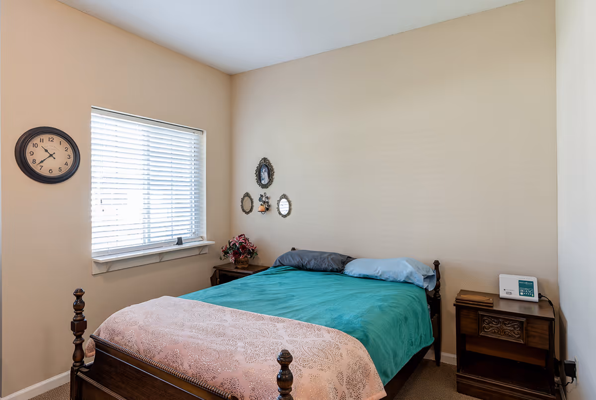 A small bedroom with a wooden bed covered with a teal blanket and a pink patterned throw. There are two pillows on the bed, one gray and one light blue. A window with white blinds is on the left wall, with a round clock above it. On either side of the bed are wooden nightstands, one with a potted plant and the other with a small electronic device. Three small decorative mirrors and a candle holder are mounted on the wall above the bed.