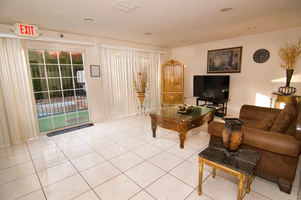 A bright living room with white tiled floors, a brown sofa with cushions, a wooden coffee table with decorative items, a small side table with a vase, a flat-screen TV on a stand, a decorative cabinet, a wall clock, and a framed painting. Large sliding glass doors with vertical blinds lead to an outdoor area with a pool and greenery. An exit sign is visible above one of the doors.