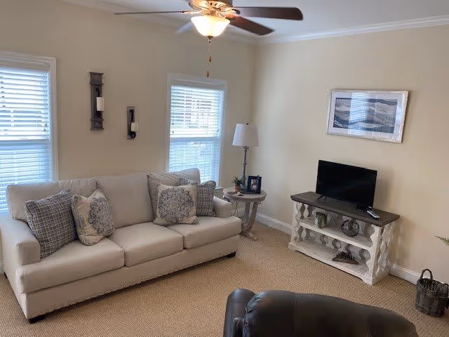 A cozy living room with a beige three-seater sofa adorned with patterned cushions, two windows with white blinds, a ceiling fan with a light, a small round side table with a lamp and picture frame, a TV on a white wooden stand, and a framed abstract artwork on the wall.