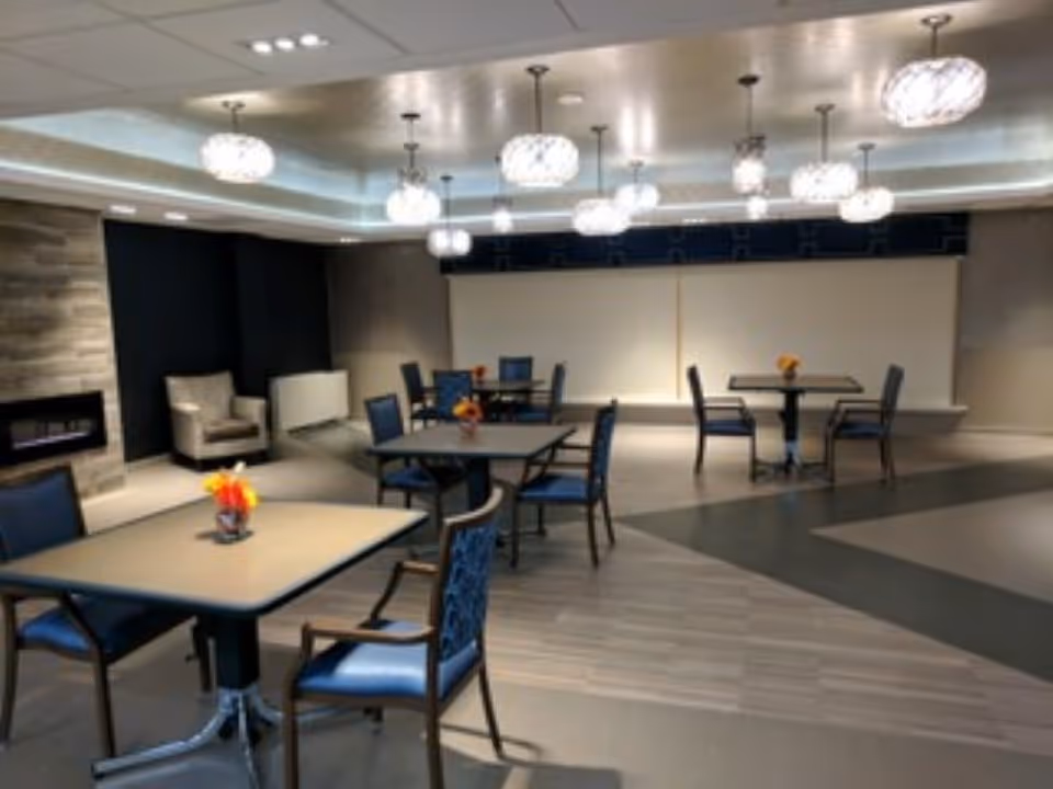 A modern dining area with several square tables and blue cushioned chairs arranged neatly. Each table has a small vase with flowers. The room features a contemporary ceiling with multiple hanging light fixtures and a cozy fireplace on the left side. There is also a comfortable armchair near the fireplace.