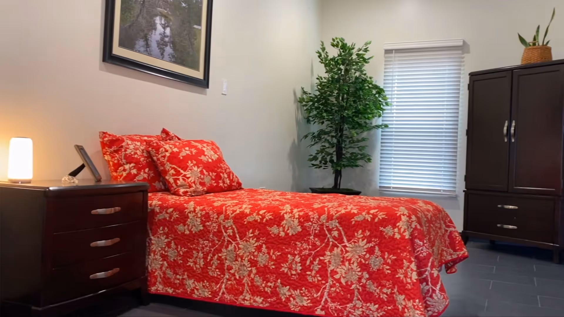 A bedroom with a red floral bedspread, a nightstand with a lamp, a potted plant by a window with blinds, and a dark wood armoire.