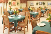A dining room with multiple wooden tables and chairs. The tables are covered with green tablecloths and set with white plates and napkins. There are flower arrangements on each table. The room has framed pictures and plants on the walls, with windows covered by curtains.