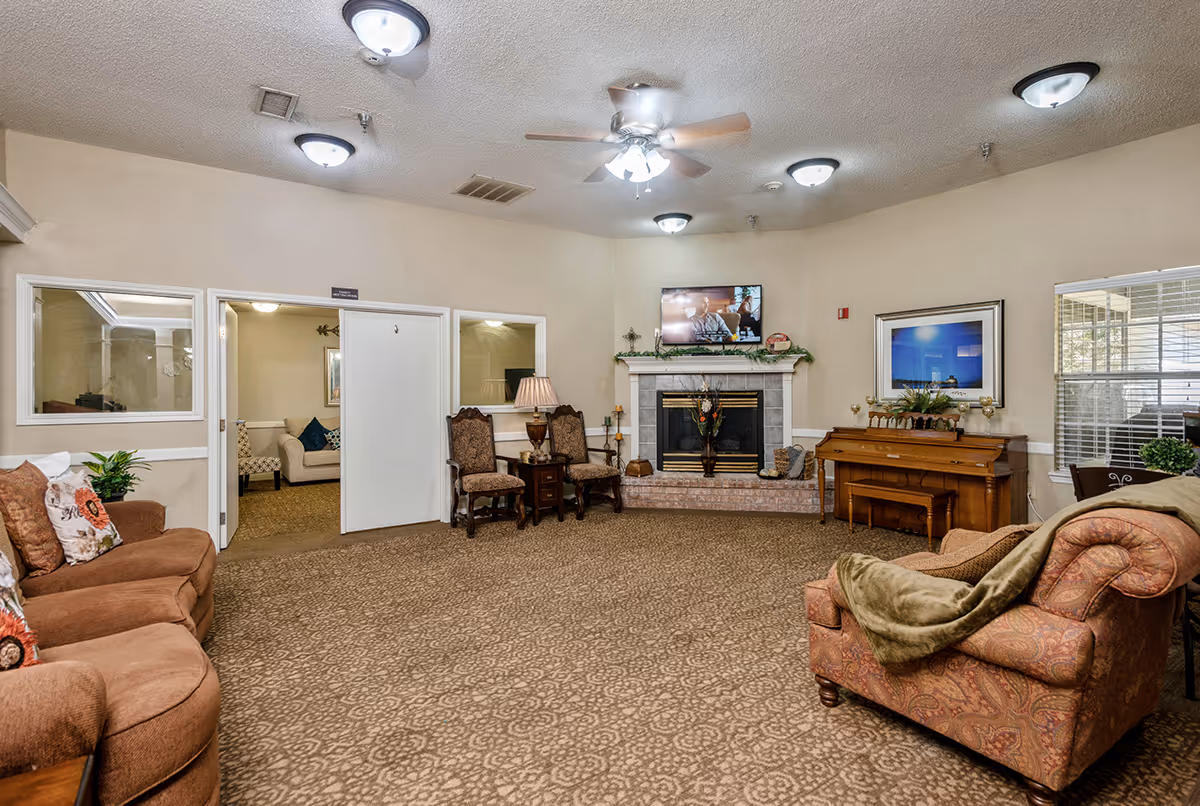 A cozy living room area in Rosefield Court featuring a patterned carpet, a fireplace with a TV mounted above it, two upholstered chairs with a small table and lamp between them, a piano with a bench, and several sofas with cushions. There are windows with blinds and ceiling lights including a ceiling fan with lights.