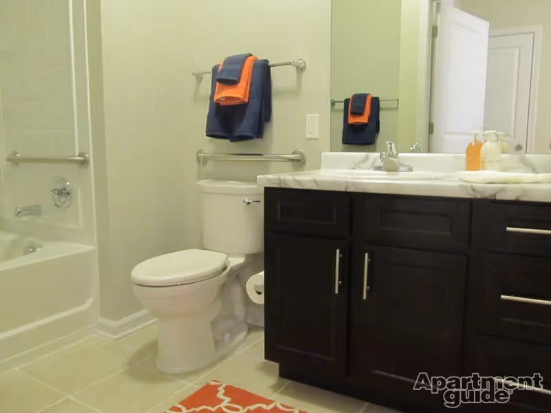 A bathroom with a white toilet, a bathtub with tiled walls, a dark wood vanity with a marble countertop, a sink with a faucet, and a large mirror. There are navy blue and orange towels hanging on a towel rack above the toilet. A red patterned rug is on the tiled floor.