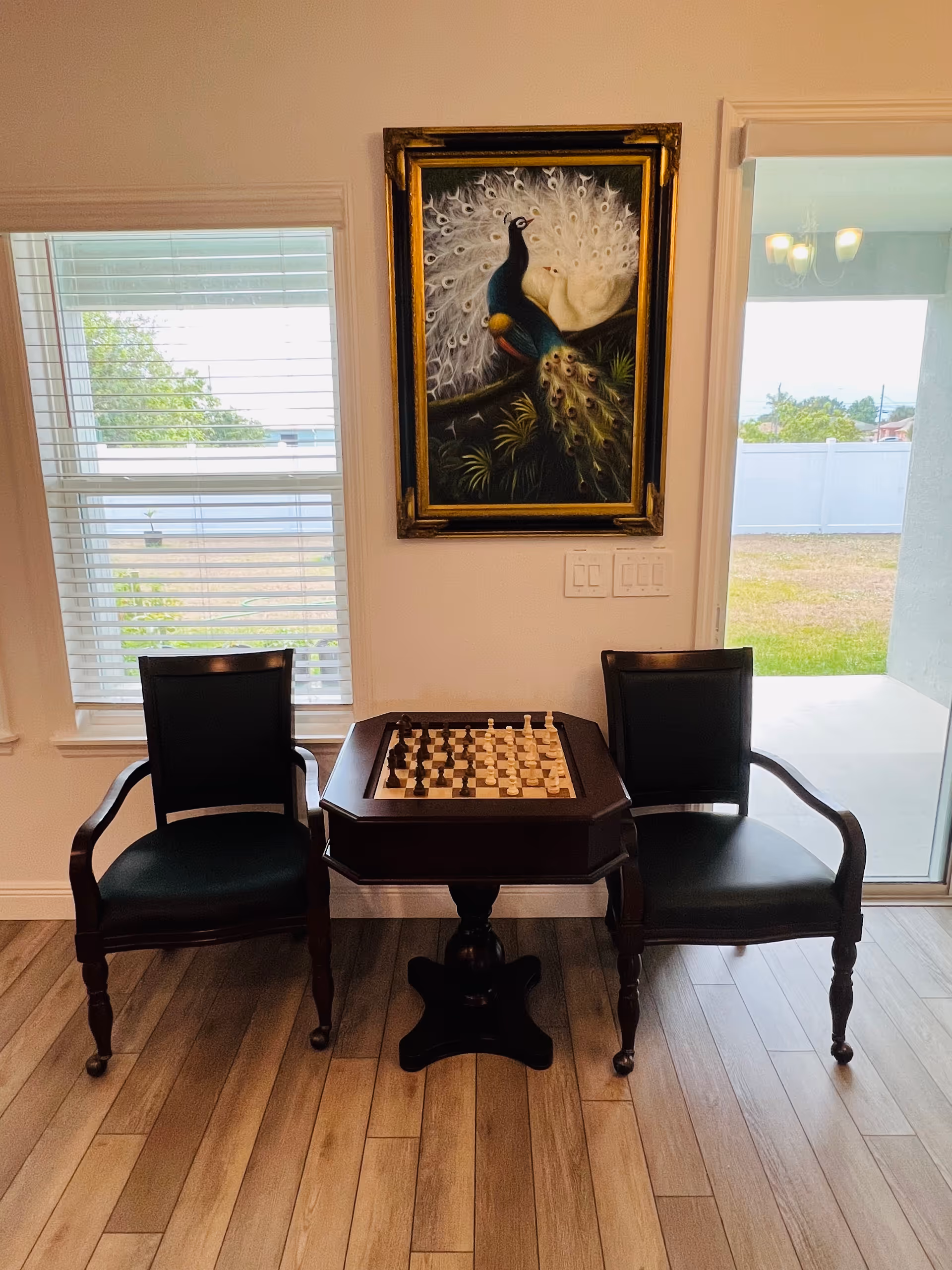 Two dark wooden armchairs flank a small chess table with a chessboard beneath a framed peacock painting, next to windows and a sliding glass door.