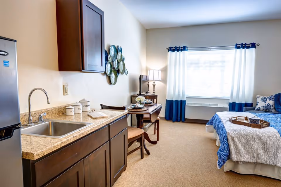 A bright and cozy studio apartment featuring a small kitchenette with a sink, countertop, and dark wooden cabinets. There is a dining table set for two with chairs, a lamp, and a TV on a stand near a large window with white and blue curtains. A bed with blue and white bedding and a tray with a cup and bowl is visible on the right side of the room.