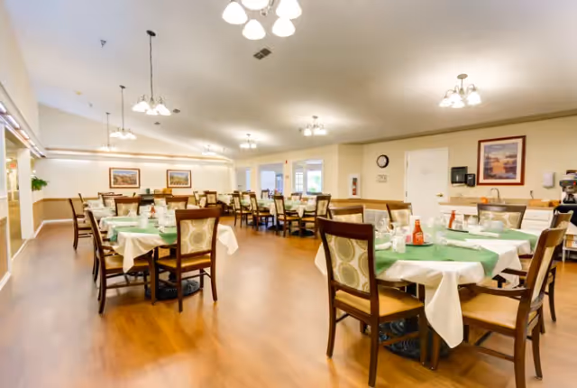 Spacious dining room with multiple round and rectangular tables set with green tablecloths and wooden chairs under ceiling lights.
