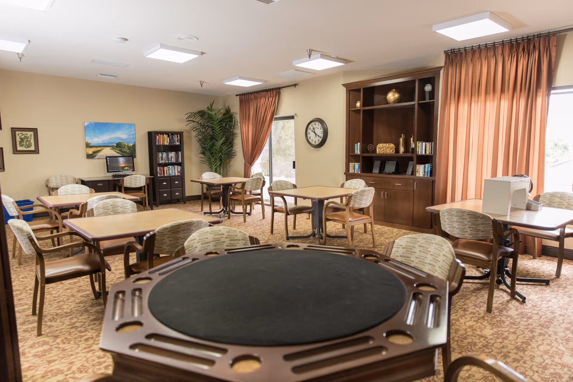 A well-lit common room with multiple tables and chairs arranged for group activities or dining. The room features a large wooden game table in the foreground, a bookshelf filled with books, a computer desk with a monitor, a large plant, wall art, a clock, and large windows with orange curtains letting in natural light.