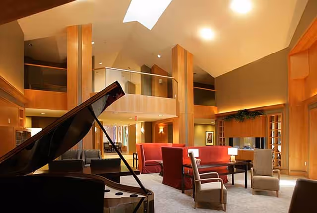 A spacious and well-lit living room area with high ceilings and wooden beams. The room features a grand piano in the foreground, red and beige upholstered chairs and sofas arranged around a coffee table, and warm lighting. There is a balcony or loft area above and decorative plants on a shelf along the wall.