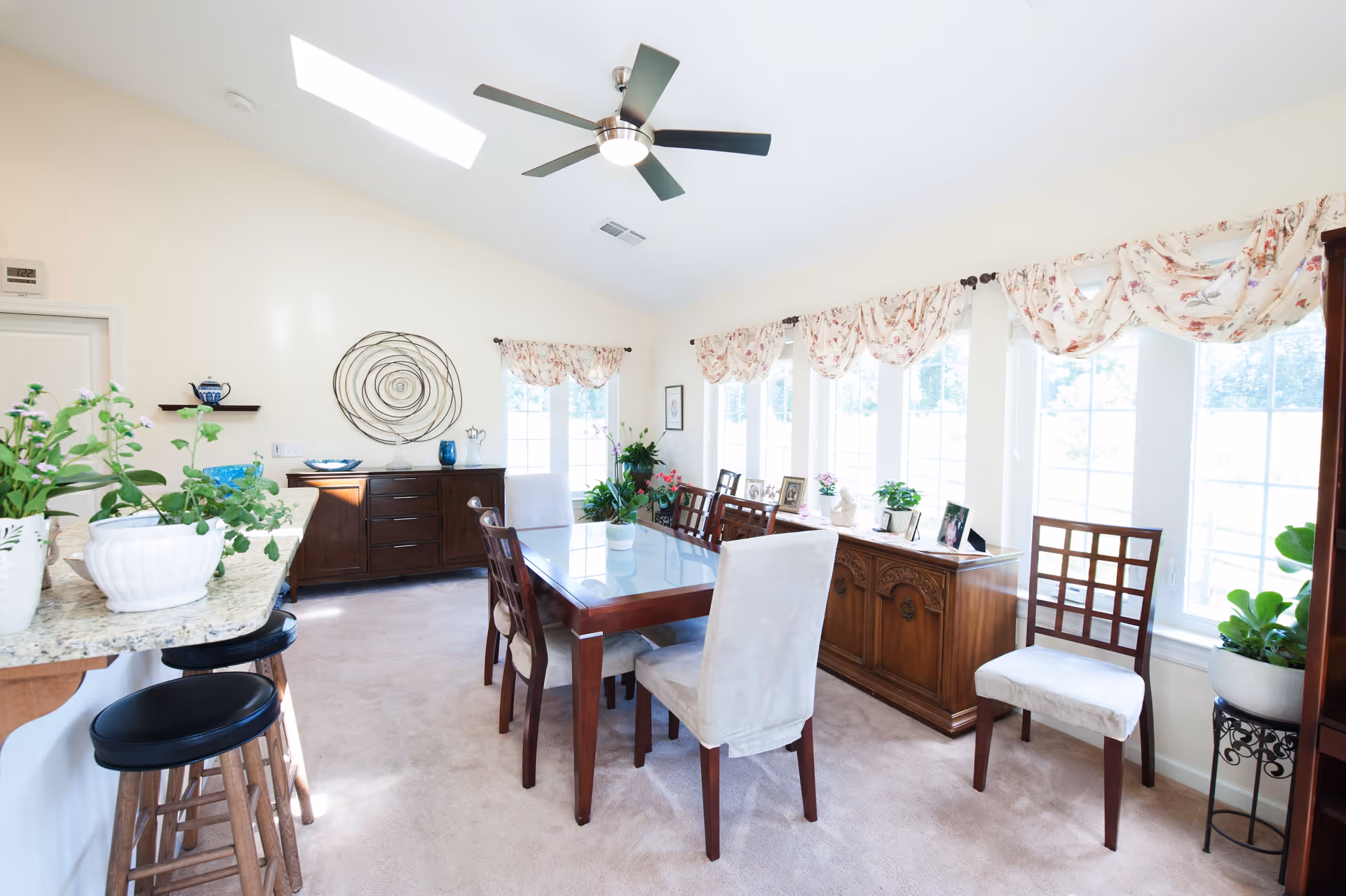 Bright dining room with a wooden table surrounded by six chairs, two of which have white slipcovers. The room features large windows with floral valances, a ceiling fan with light, and a sideboard with framed photos and plants. There is a kitchen counter with bar stools and potted plants on the left side.