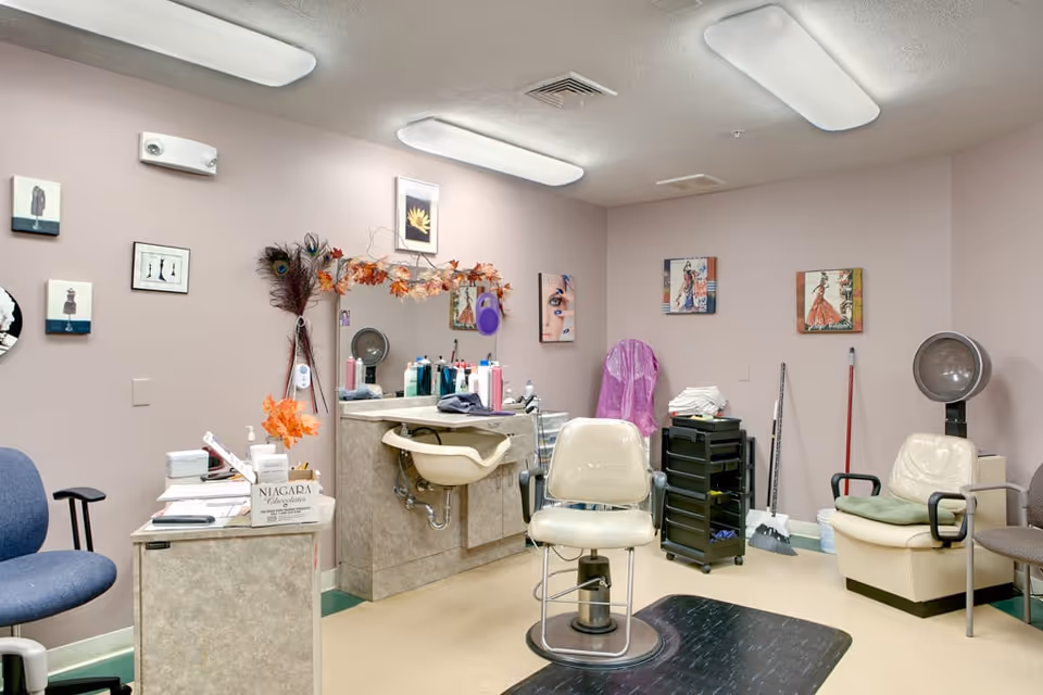Interior of a salon room with a salon chair in the center, a sink with various hair care products on the counter, a mirror decorated with autumn leaves, and several chairs including a salon dryer chair. The walls are adorned with framed pictures and the room is lit by ceiling lights.