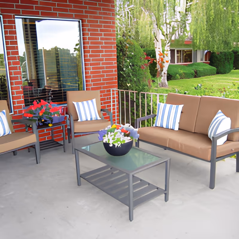 Outdoor patio with brown cushioned seating, a glass-top coffee table holding a bowl of flowers, potted plants, and a brick wall overlooking a green lawn.