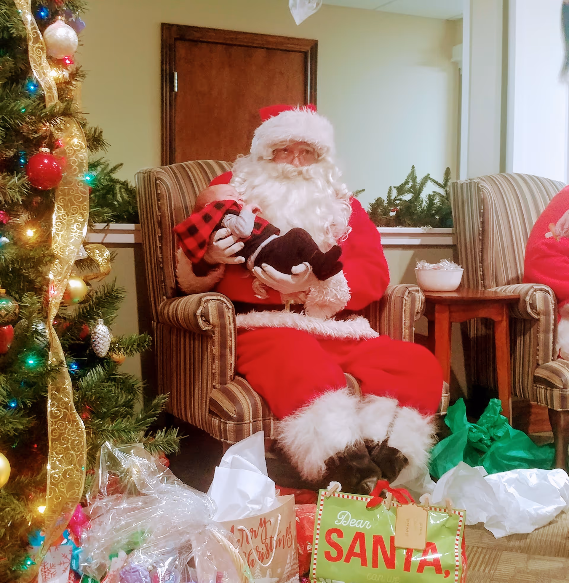 A person dressed as Santa Claus sitting in a striped armchair holding a baby dressed in a red and black plaid outfit. To the left is a decorated Christmas tree with lights, ornaments, and gold ribbon. In front of Santa are wrapped gifts and gift bags, including one with the text 'Dear Santa'. The setting appears to be a cozy indoor room with holiday decorations.