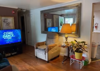 A cozy living room area with a beige recliner chair next to a small side table holding a lamp and some items. A large decorative mirror hangs on the wall above the chair, reflecting part of the room. A flat-screen TV is on the left side displaying a show called 'Come Dance With Me.' There is a plant in a yellow pot on a small stand near the chair. The room has wooden flooring and light-colored walls.