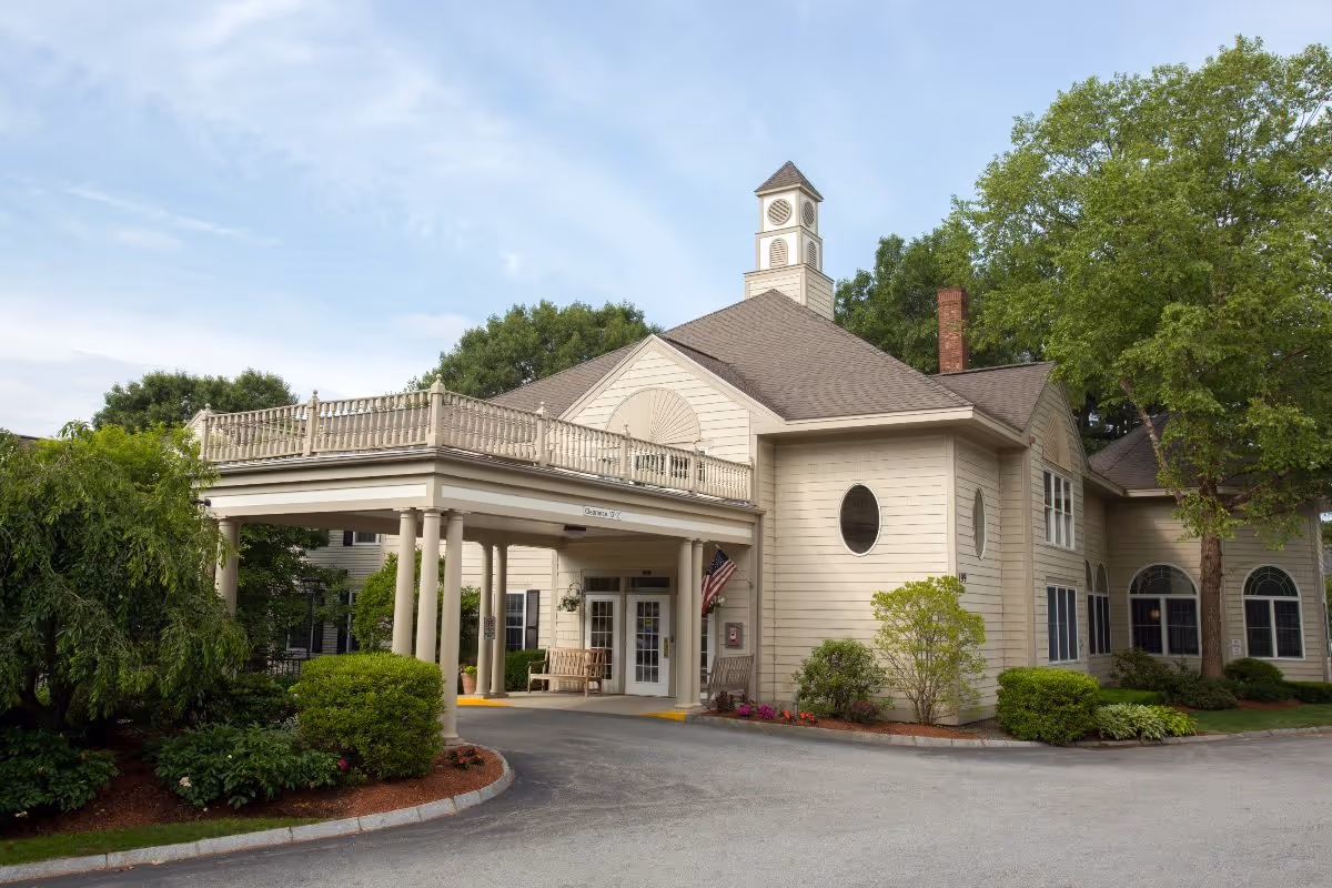 Exterior view of a senior living facility building with beige siding, a covered entrance supported by columns, an American flag, and surrounding greenery including trees and bushes under a partly cloudy sky.