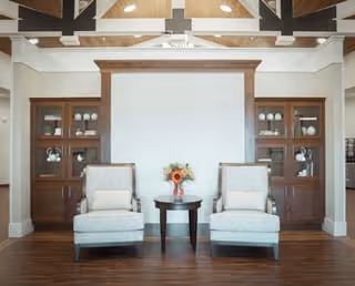 A cozy seating area with two upholstered armchairs facing forward, separated by a small round wooden table with a vase of flowers. Behind the chairs is a built-in wooden cabinet with glass doors displaying white dishes and decorative items. The room features a high ceiling with exposed beams and a ceiling fan.