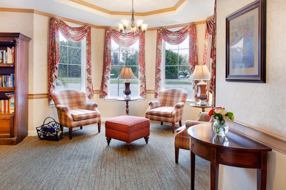 Cozy sitting area with plaid armchairs, an upholstered ottoman, side tables with lamps, a bookshelf, and bay windows with patterned curtains.