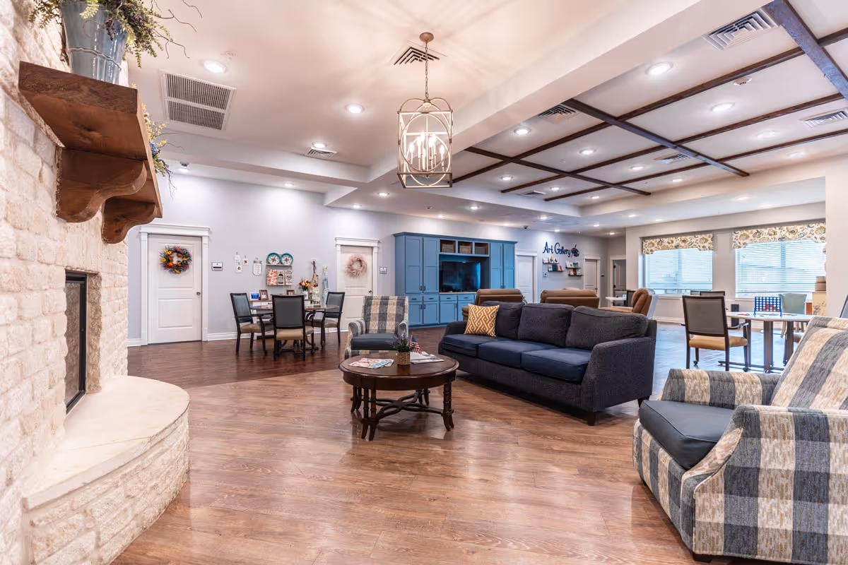 A spacious and well-lit assisted living common area featuring a stone fireplace on the left, a dark blue sofa with a yellow patterned pillow, two plaid armchairs, a round wooden coffee table, and a dining table with chairs in the background. The room has wooden flooring, recessed lighting, a decorative ceiling with wooden beams, and large windows with floral valances letting in natural light. A blue entertainment center with a TV and an 'Art Gallery' sign is visible in the back.