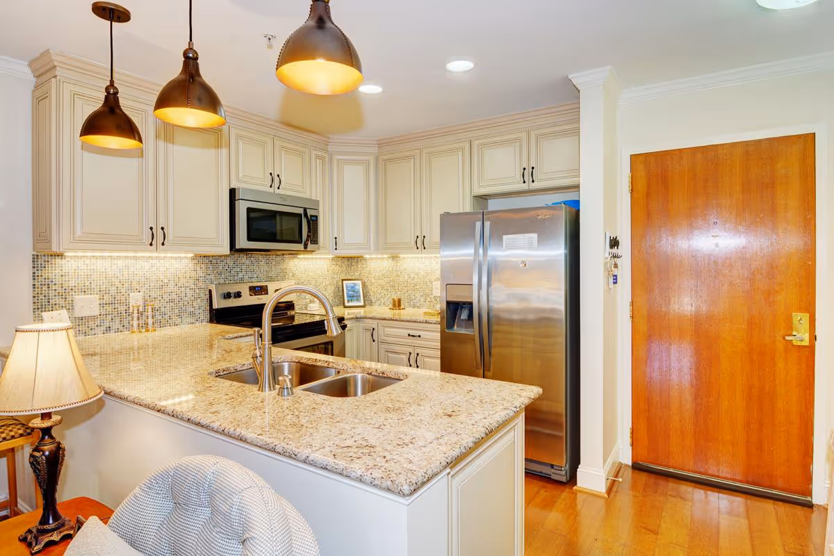 Bright modern kitchen with a granite island and double sink, pendant lights, stainless steel refrigerator and microwave, and a wooden entry door.