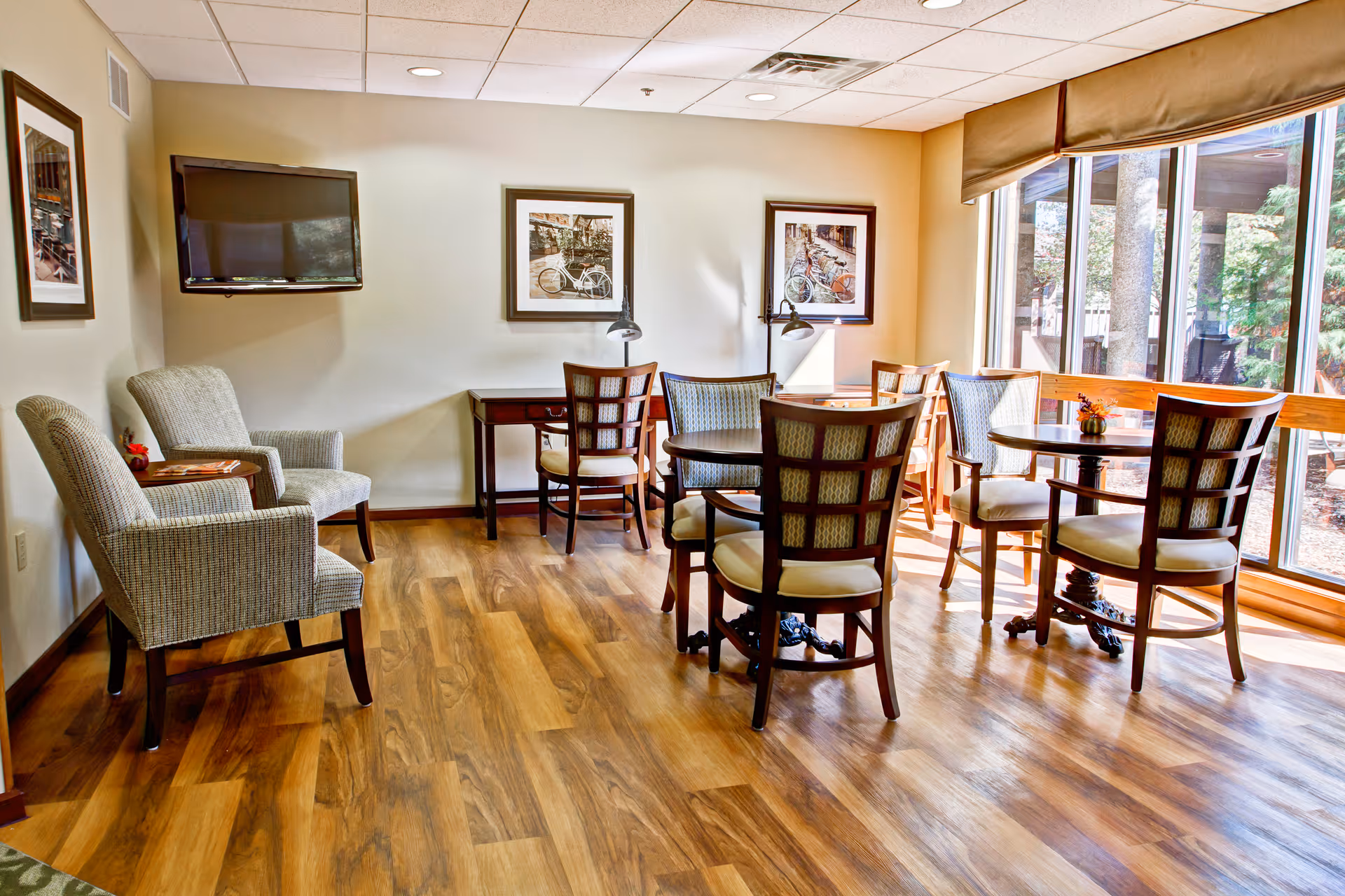 Sunny common room with round tables and chairs, armchairs, a wall-mounted TV, and large windows overlooking the courtyard.