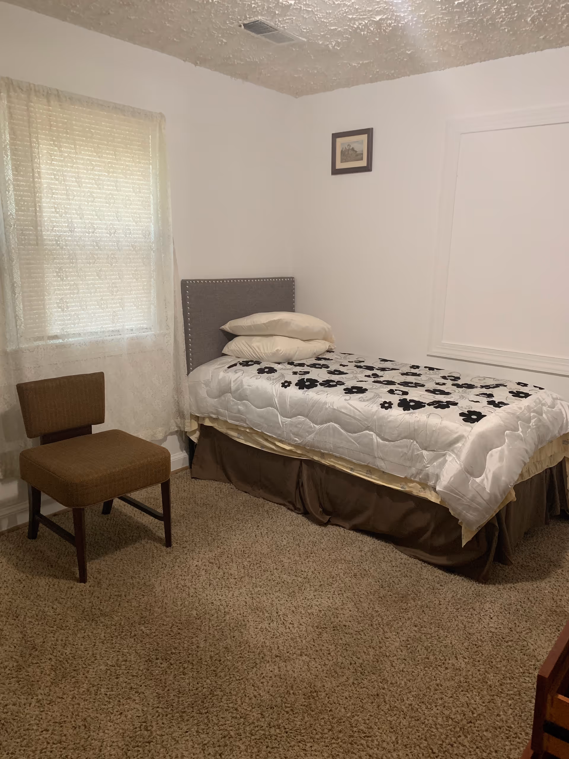 A small bedroom with a single bed covered with a white and black floral patterned comforter and two pillows. There is a brown chair next to the bed, a window with sheer curtains, beige carpet flooring, and a small framed picture on the white wall.