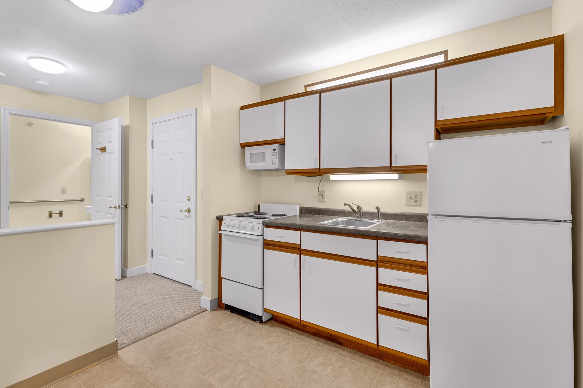 A small kitchen area in a senior living facility featuring white cabinets with wooden trim, a white refrigerator, a white stove with an oven, a microwave mounted above the stove, and a sink with a countertop. The kitchen has beige walls and a tiled floor. In the background, there is a hallway with two white doors and a carpeted floor.
