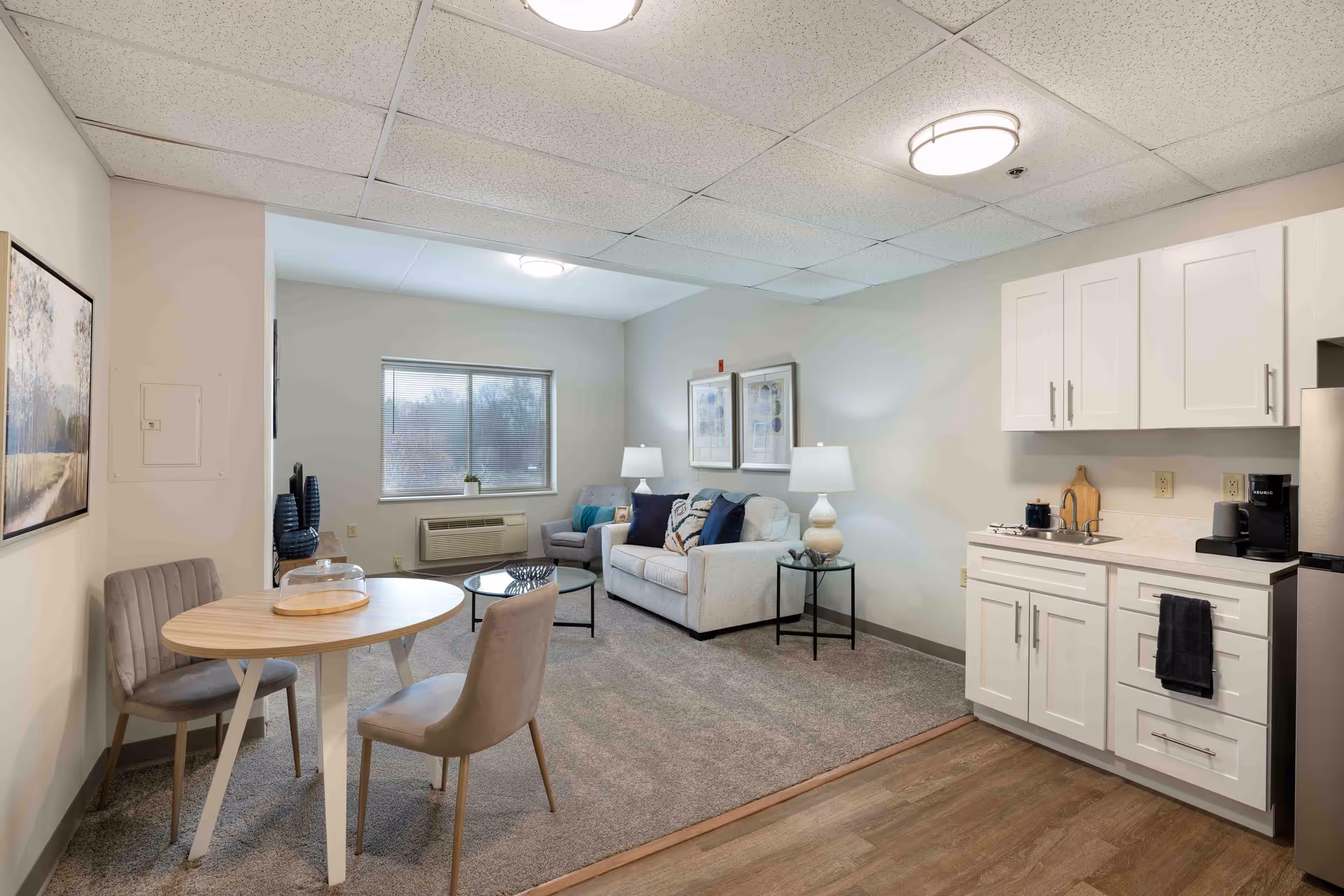 A cozy senior living community interior featuring a small dining area with a round wooden table and two upholstered chairs, a living room with a white sofa, armchair, coffee table, and two lamps, and a kitchenette with white cabinets, a sink, coffee maker, and a stainless steel refrigerator. The room has a window with a view outside and neutral-colored walls and carpet.