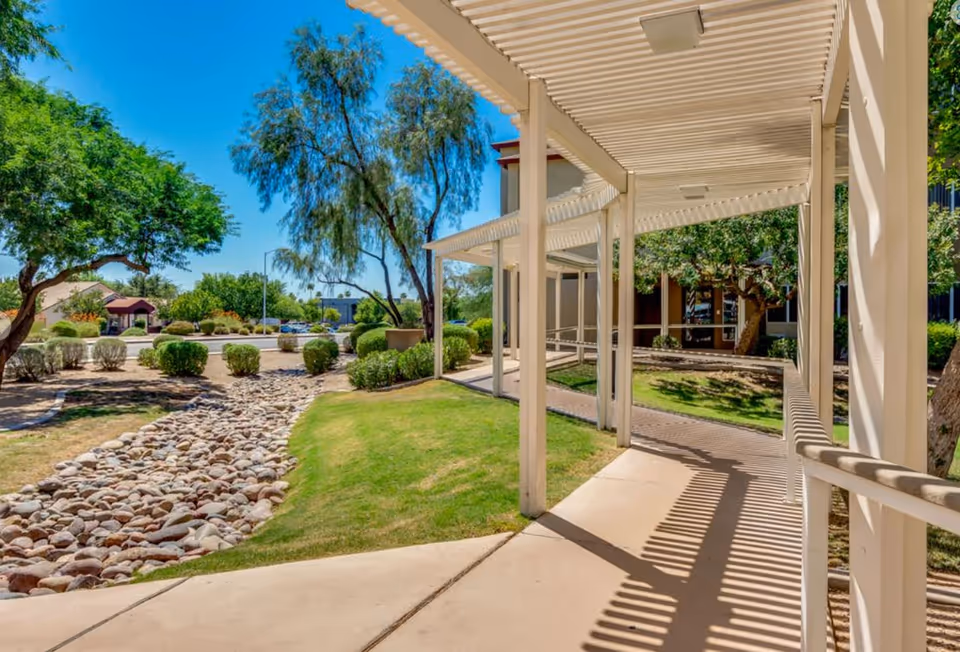 Outdoor walkway with white pergola structure casting shadows on the concrete path, surrounded by green grass, bushes, trees, and a dry rock stream bed under a clear blue sky.