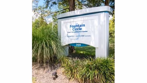 Outdoor landscaped sign reading "Fountain Circle Care & Rehabilitation Center" with trees and grasses around it.