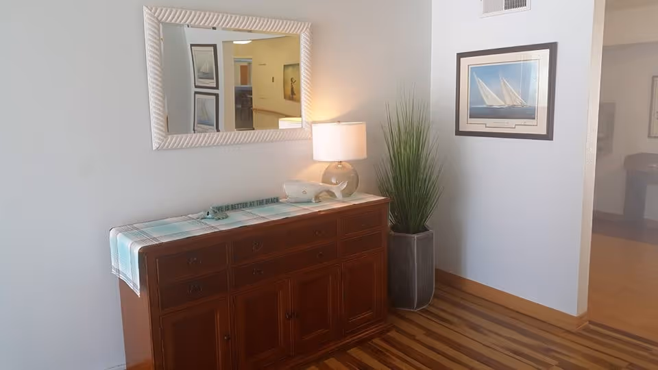 Interior corner of a room with a wooden cabinet topped with a blue and white plaid runner, a glass lamp, a decorative whale, and a small sign. Above the cabinet is a large white-framed mirror reflecting part of the hallway. To the right of the cabinet is a tall green plant in a gray pot, and a framed picture of sailboats hangs on the wall. The floor is wooden with a striped pattern.