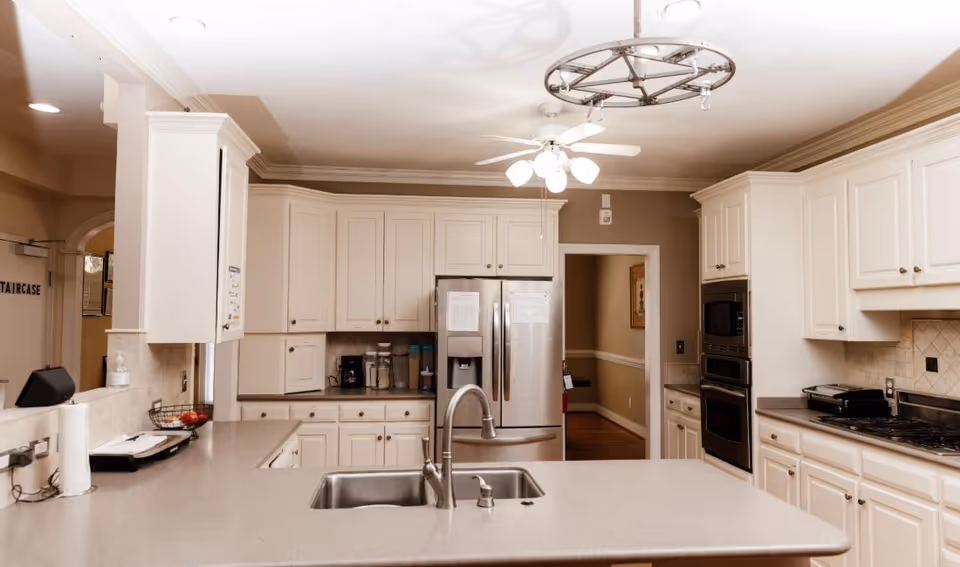 A spacious kitchen with white cabinetry, a large island with a double sink and faucet, stainless steel refrigerator, built-in oven and microwave, a gas stove, and a ceiling fan with lights. The kitchen has beige walls and a tiled backsplash.