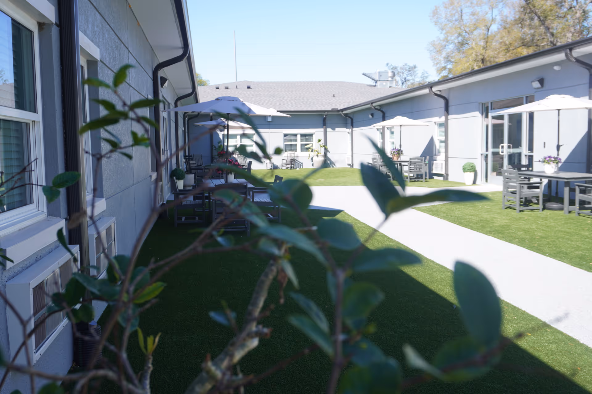 Outdoor courtyard with a grassy lawn, paved walkway, patio tables and umbrellas alongside a single-story building.
