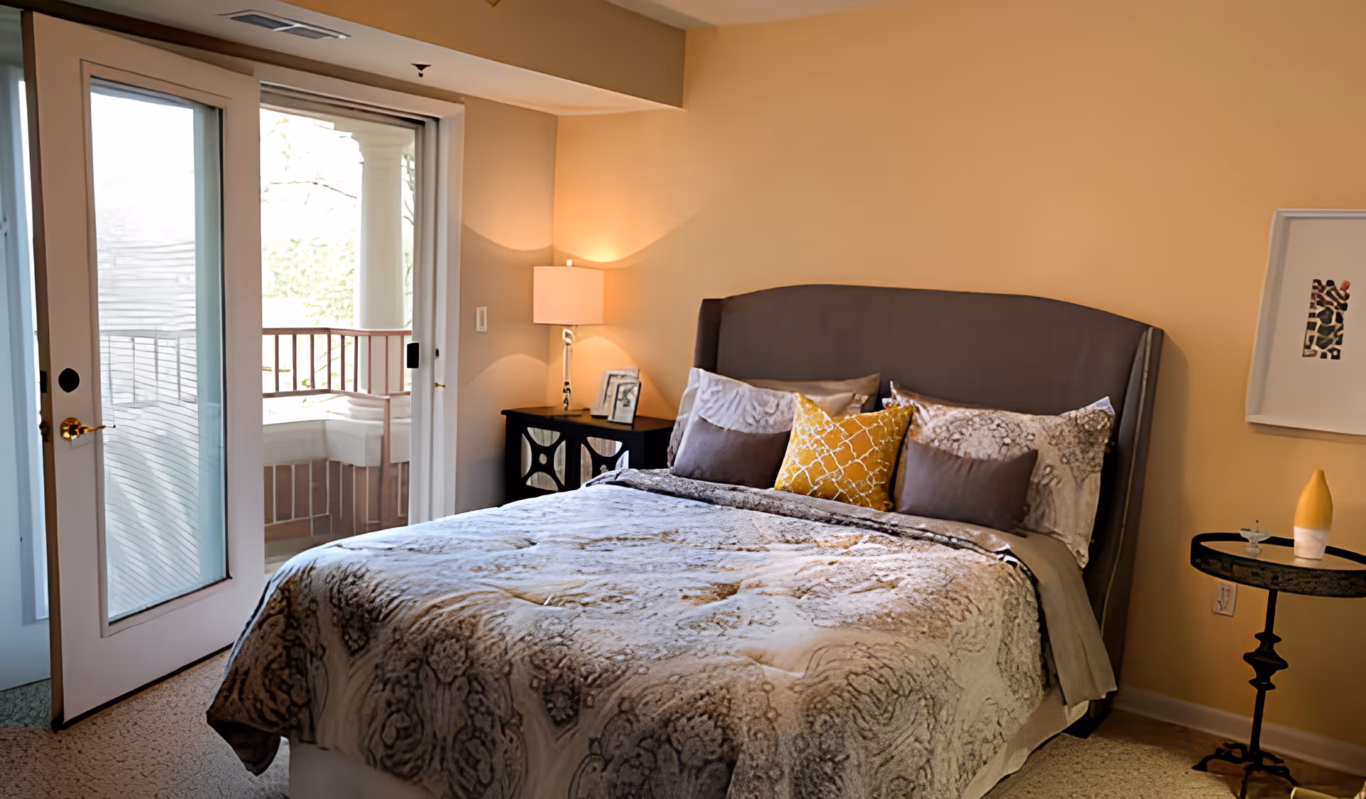 A cozy bedroom with a large bed featuring a patterned comforter and multiple pillows, including a yellow decorative pillow. There is a bedside table with a lamp and framed photos on the left side, and a small round table with a decorative item on the right. The room has beige walls and a sliding glass door leading to a balcony with white columns and a railing.