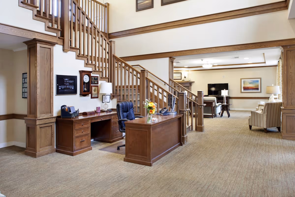 Interior view of a senior living facility featuring a wooden reception desk with a black office chair, a vase with flowers, and a telephone. Behind the desk is a wooden staircase with railings leading to an upper floor. The room has beige walls and carpeted floors. In the background, there is a seating area with armchairs, a television, a fireplace, and framed artwork on the walls.