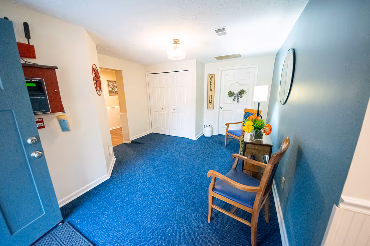 An entryway area with blue carpet and light blue walls. There are two wooden chairs with blue cushions, a small table with a lamp and flower arrangements, a round mirror on the wall, and a door with a wreath. A welcome sign is mounted on the wall next to the door. The open blue entrance door reveals a fire alarm panel on the wall.