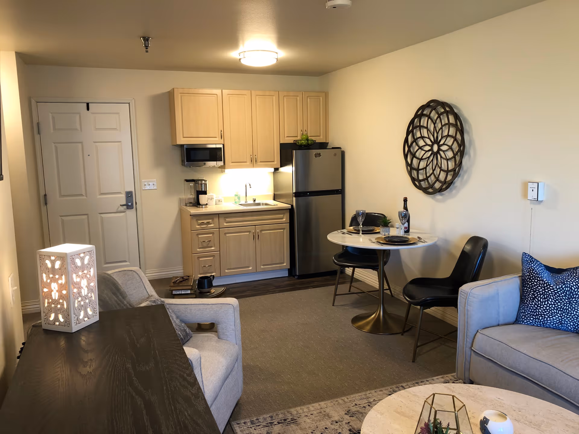 A cozy living area in a senior living facility featuring a small kitchenette with light wood cabinets, a microwave, a coffee maker, and a stainless steel refrigerator. There is a round dining table set for two with black chairs, a decorative wall piece above the table, and a comfortable seating area with a gray armchair and sofa with blue patterned pillows. A decorative lamp sits on a dark wooden table next to the armchair.