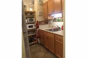 Small kitchen with wooden cabinets, a sink under a window, a microwave and toaster oven on shelves, and various kitchen items on the counter and shelves.