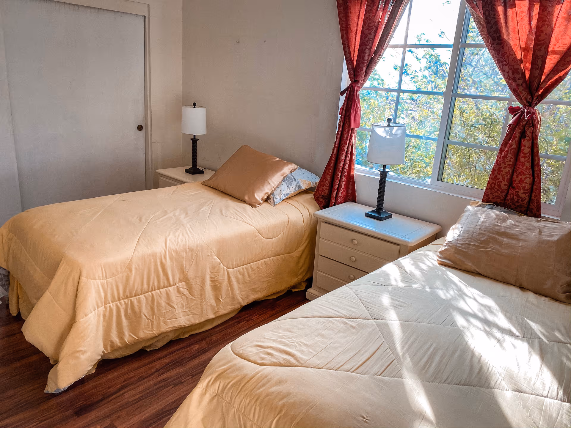 A bedroom with two single beds covered in beige comforters and pillows. Between the beds is a white nightstand with a lamp on it. The room has wooden flooring, a window with red curtains letting in sunlight, and a closed white door on the left wall.