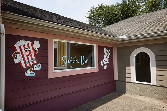 Exterior wall of a building with a window reading 'Snack Bar' and painted cartoon popcorn and soda characters.