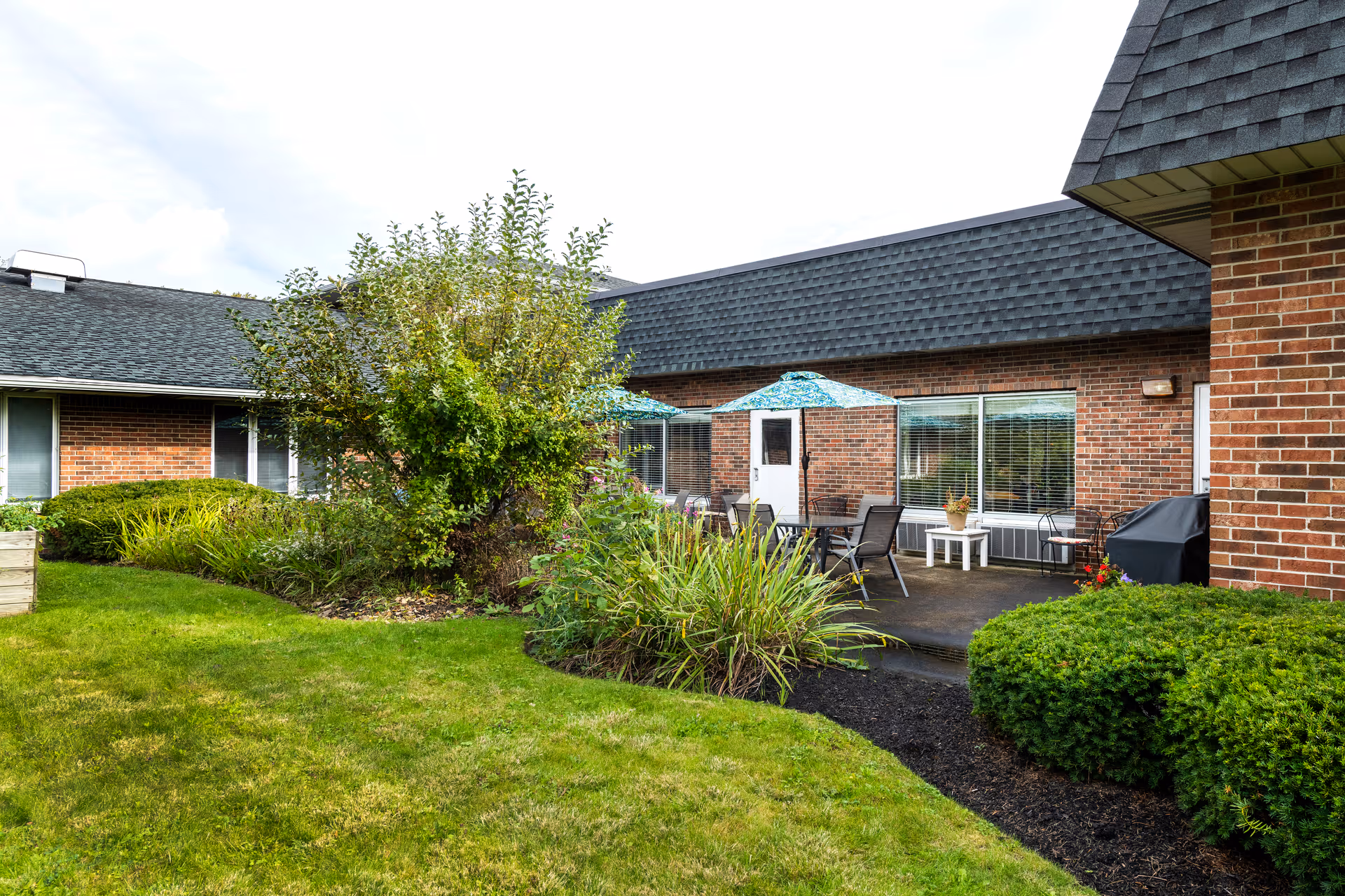 Outdoor patio area at Elderwood Village at Bassett Park with green grass, bushes, a tree, and a brick building in the background. The patio has tables, chairs, and umbrellas.