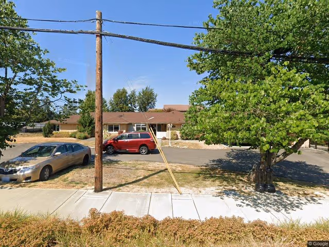 Exterior view of a single-story residential building with a brown roof and beige walls, surrounded by trees and parked cars on a sunny day. A sidewalk and a utility pole with overhead wires are visible in the foreground.