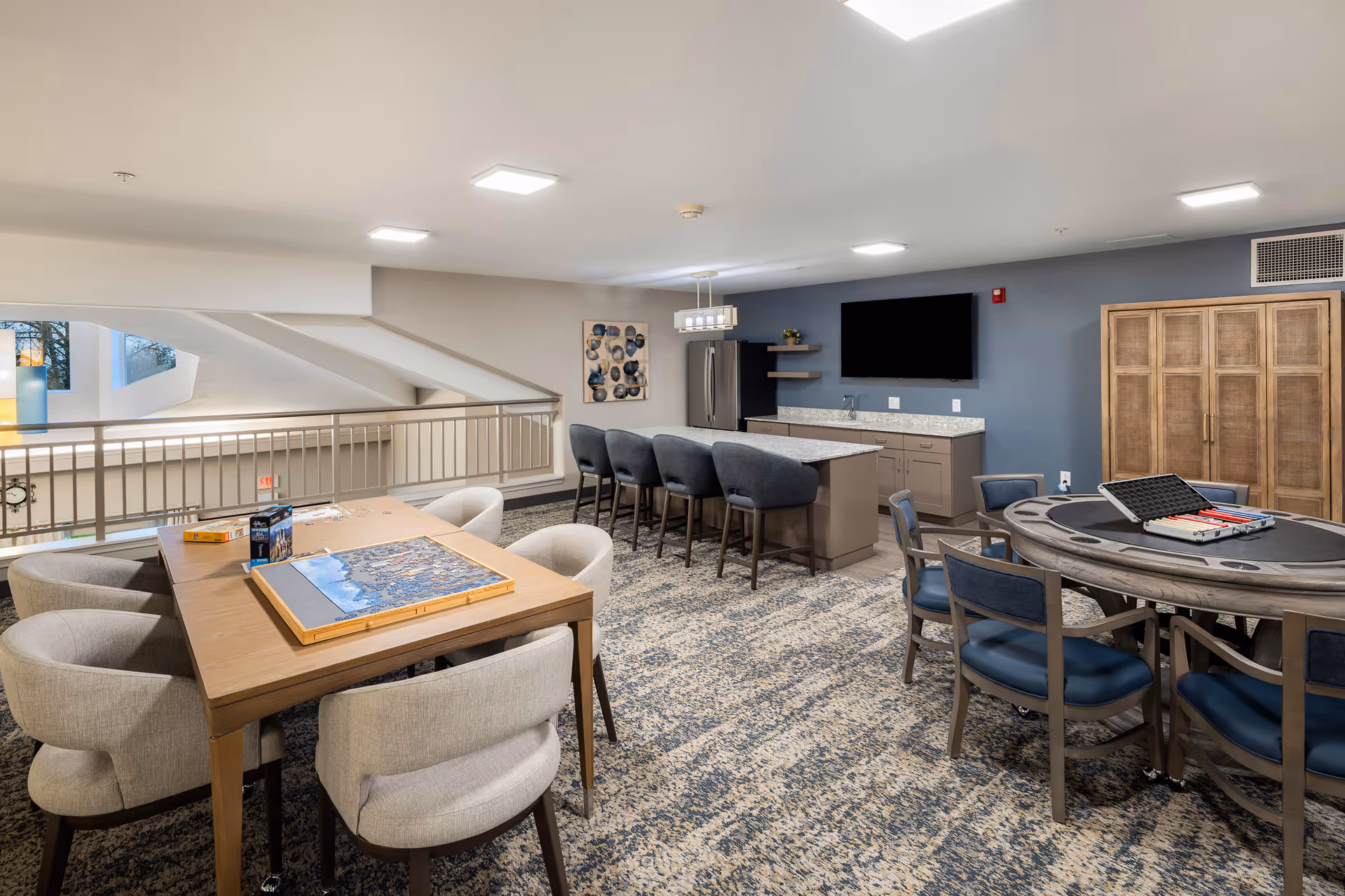 A spacious senior living community common area with a large wooden table and cushioned chairs on the left, featuring a partially completed puzzle. To the right, there is a round table with chairs and a board game on it. In the background, there is a kitchen area with a countertop, bar stools, a refrigerator, and a wall-mounted TV. The room has a carpeted floor and modern lighting.