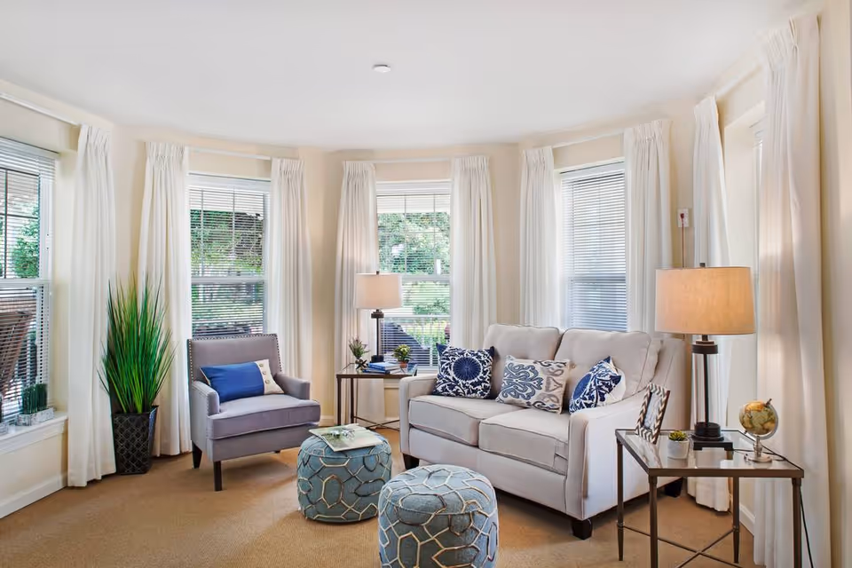 A bright living room with beige walls and carpet, featuring a light gray sofa with patterned blue and beige cushions, a gray armchair with a blue pillow, two blue patterned ottomans, two side tables with lamps, a small globe, and a tall green plant near the windows with white curtains.